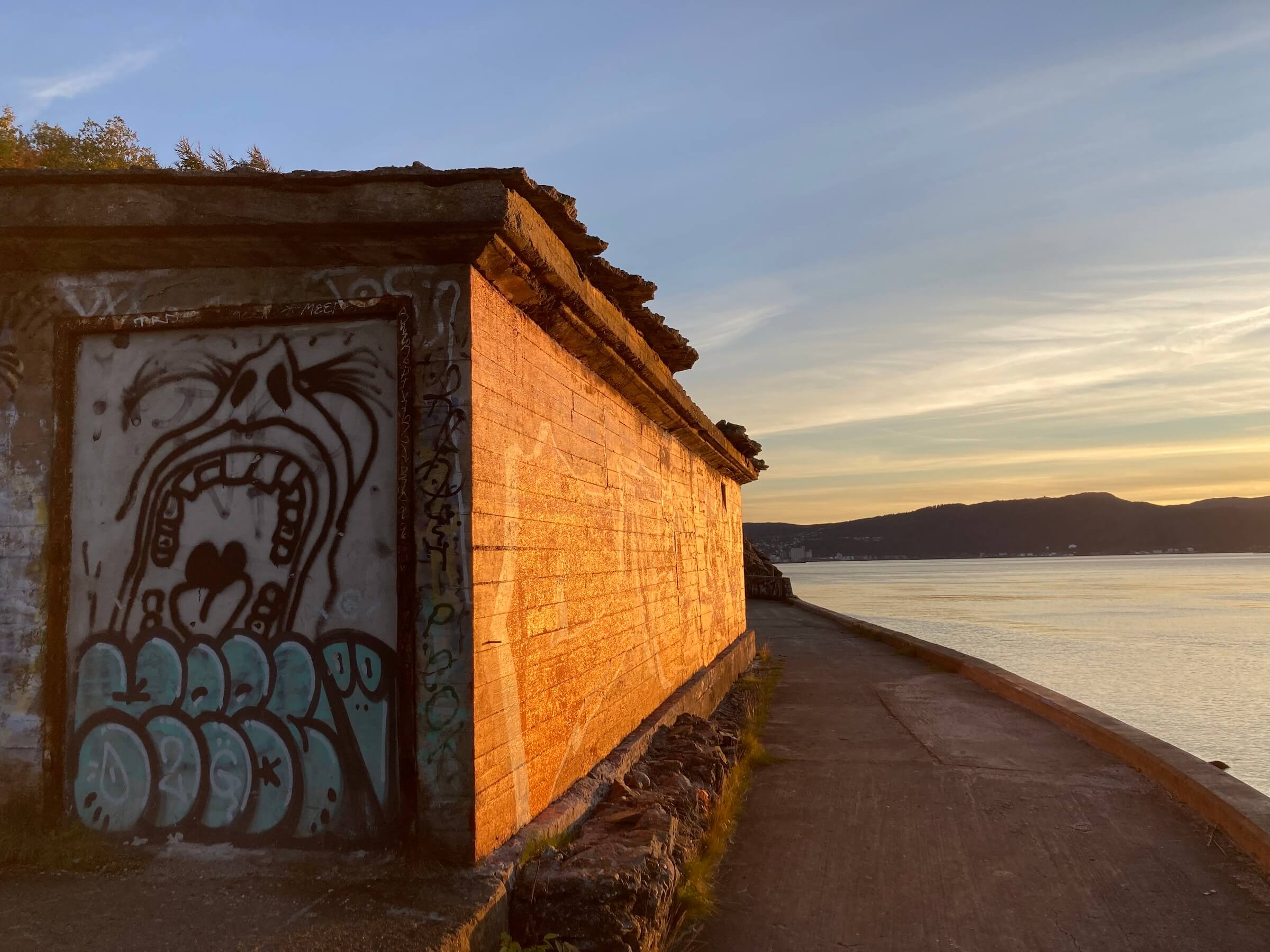 On the left side an old derelict bunker with graffiti of a face, on the right side sunset over Trondheimsfjorden.